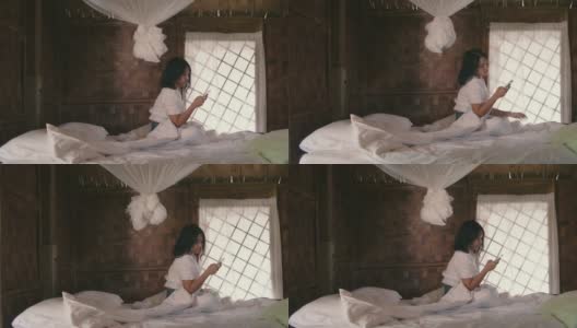 年轻女子坐在卧室白色的床上用智能手机，享受微风吹拂她的头发。高清在线视频素材下载