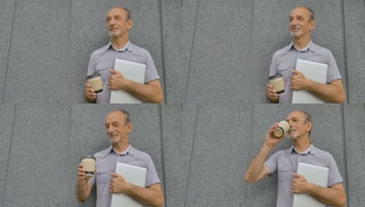 男人的肖像年长的同事与银色笔记本电脑和一杯咖啡等待会议在深灰色的墙壁在早上，远程工作，咖啡休息高清在线视频素材下载