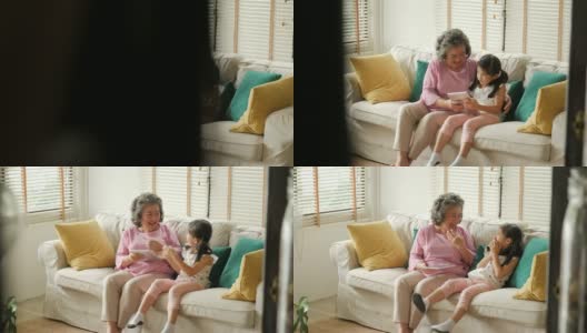 一名亚洲资深女性和一名年轻女孩坐在家里的沙发上，用数码平板电脑聊天和微笑。有科技观念的人。高清在线视频素材下载