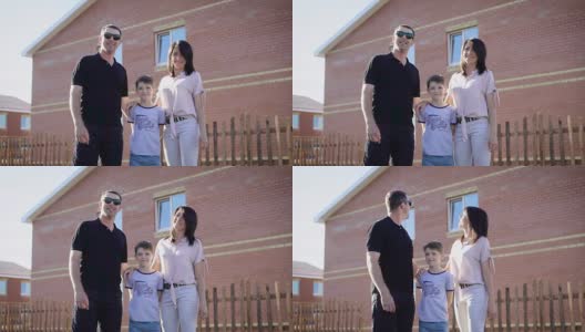 阳光明媚的夏日里，一家人穿着休闲的衣服站在新家附近。父亲，母亲和儿子走在建筑外面，背景是建筑高清在线视频素材下载