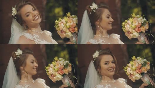 年轻美丽的新娘带着婚礼花束，微笑着待在古老的石墙附近高清在线视频素材下载