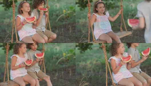 快乐的女孩和男孩坐在秋千上吃着西瓜，夏天的照片。孩子们拿着西瓜高清在线视频素材下载