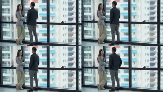 快乐的亚洲商人和女商人讨论商业战略在一个高楼大厦的现代办公室城市景观的背景。两个工人在城市摩天大楼的窗前交谈高清在线视频素材下载