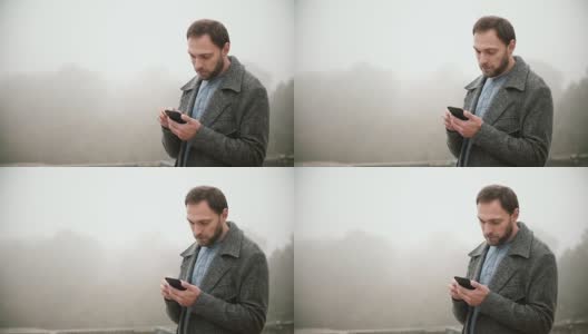 年轻英俊的男子站在雾蒙蒙的清晨公园的肖像。胡须男使用带触摸屏的智能手机高清在线视频素材下载
