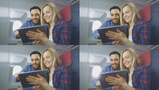 在商用飞机的一个板上，漂亮的年轻金发美女和英俊的西班牙男性使用平板电脑和微笑。阳光透过飞机窗户。高清在线视频素材下载