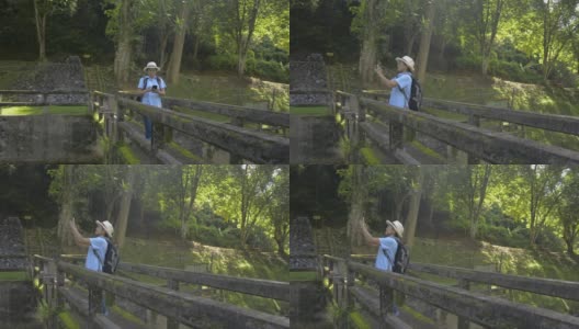 亚洲女游客戴着草帽和背包走在旧混凝土桥上，用手机拍照。游客喜欢在夏日的阳光下在森林公园里度过时光。高清在线视频素材下载