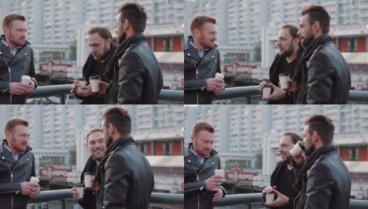 三个留着胡子的英俊青年在城市的一座桥栏杆附近聊天，边喝咖啡边聊天。缓慢的莫高清在线视频素材下载