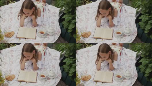 年轻女子躺在夏天的草地上看书，地毯上放着饼干和茶。浪漫的女人拿着书、茶和饼干在夏日的花丛中放松。高清在线视频素材下载