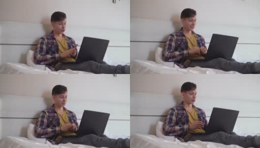 年轻的白人男子坐在家里的床上，穿着便服，用他的笔记本电脑视频聊天。C高清在线视频素材下载