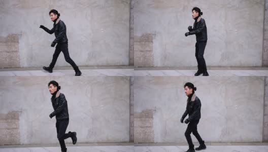 一名留着小胡子的中国青年在城市混凝土墙旁的街道上跳舞。他穿着夹克，看起来有点笨拙，4k镜头，慢动作。高清在线视频素材下载