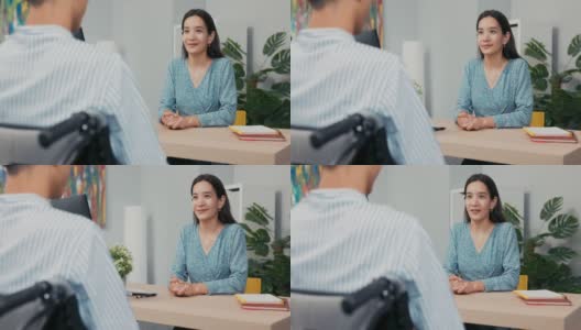 一个坐着轮椅的残疾人面试，一个亚裔韩国美女向穿着衬衫的男孩提问，她记下了答案，他们正坐在公司办公室的桌子两边高清在线视频素材下载