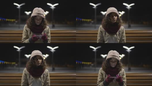 美丽的女孩独自坐在晚上的公交车站，浏览着小玩意上的日程安排高清在线视频素材下载