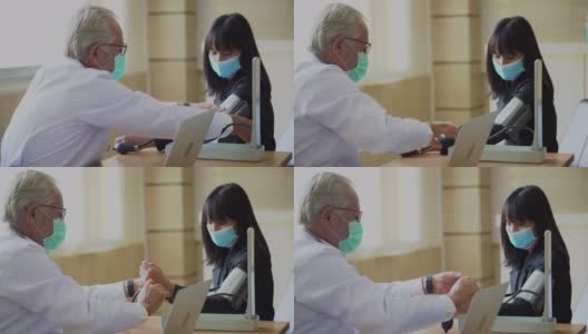 资深医生戴着外科口罩在医院测量血压，用笔记本电脑与年轻的亚洲病人交谈。新冠肺炎大流行或疫情期间的新常态高清在线视频素材下载