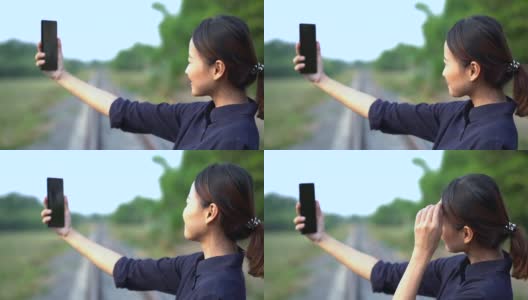 年轻美丽的亚洲人把头发绑在后面，用智能手机自拍，背景是模糊的铁路和森林高清在线视频素材下载