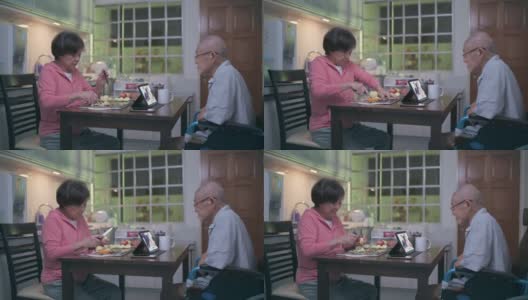 一位坐着轮椅、戴着气管造口管的亚洲老人，在晚餐后的闲暇时间，妻子在厨房递给他苹果时，他用数码平板电脑给家人打电话高清在线视频素材下载