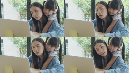 一位亚洲母亲带着她可爱的小女儿在家工作。当妈妈在笔记本电脑上工作时，一个亚洲小孩从后面拥抱她的妈妈。高清在线视频素材下载