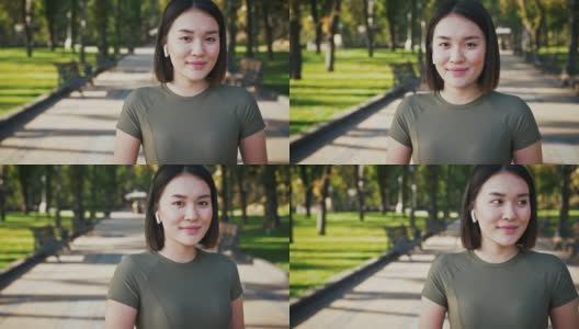 可爱的亚洲女人戴着无线耳机在城市公园对着摄像头微笑，跟踪拍摄高清在线视频素材下载
