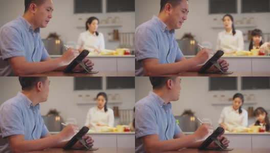 亚洲父亲写便条和工作平板电脑等待他的妻子和女儿准备晚餐的食物。他很忙，在新冠肺炎大流行期间不得不在家工作。新标准的概念。高清在线视频素材下载