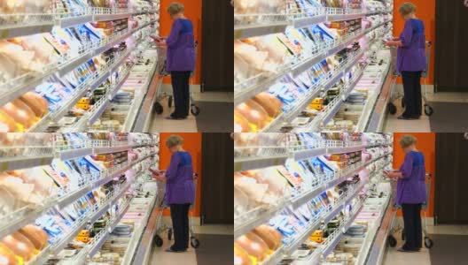 老妇人推着购物车在超市的冷藏区购买乳制品或冷藏食品。奶奶呆在商店冰箱附近，从冰箱里拿东西高清在线视频素材下载