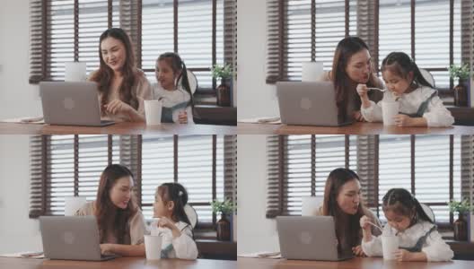 自由职业者亚洲母亲工作在桌子上与她的笔记本电脑，她的女儿吃面条在客厅在家里，幸福的家庭人的生活方式休闲活动概念。高清在线视频素材下载