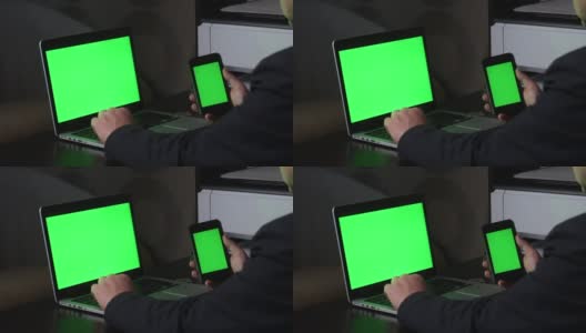 一名男子在一台绿色屏幕的笔记本电脑前工作。他手里拿着一部绿色屏幕的智能手机。附近有一枚加密货币。Chromakey。高清在线视频素材下载