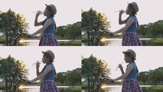 日落时分，一个戴着帽子的小女孩站在河边的公园里，骑着自行车，从一个透明的瓶子里喝水。高清在线视频素材下载