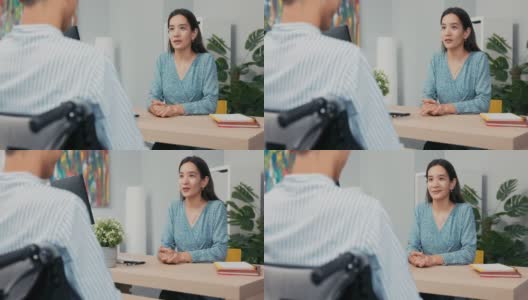 一个坐着轮椅的残疾人面试，一个亚裔韩国美女向穿着衬衫的男孩提问，她记下了答案，他们正坐在公司办公室的桌子两边高清在线视频素材下载
