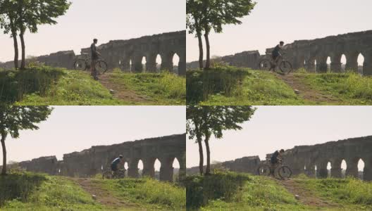 骑自行车的人在雄伟的古罗马渡槽前的土路上骑着自行车上山。年轻有吸引力的运动员与蓝色t恤和短裤运动装在parco degli acququedotti在罗马高清在线视频素材下载