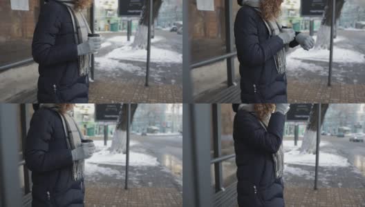 在冬天的一天，一个陌生的年轻女子穿着暖和的衣服在车站等车，喝着咖啡，看着手表。高清在线视频素材下载