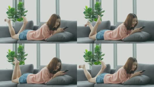 年轻美丽的亚洲女人使用智能手机与幸福，而躺在沙发上与城市的看法，人们在电话，生活方式高清在线视频素材下载