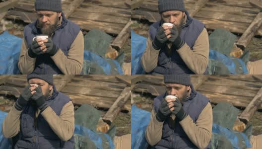 一个无家可归的人在废墟附近喝热茶，在疫情期间帮助贫困和饥饿的人们高清在线视频素材下载