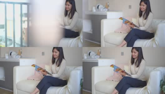 年轻的亚洲女人坐在家里的沙发上看杂志高清在线视频素材下载