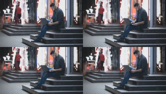 晚上，一个留着胡子的英俊男子坐在拥挤的街道上，把笔记本电脑放在腿上打字。自由工作的概念。聊天、发电子邮件、搜索或约会。高清在线视频素材下载