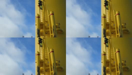 低角度查看风力发电机和绳索进出技术员(手工高工人，工业攀登者)爬上梯子。戏剧性的天空和身后旋转的叶片。相机倾斜。高清在线视频素材下载