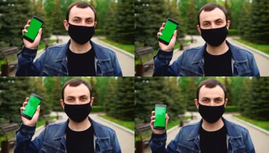身着黑色医用口罩的男子在户外展示绿色屏幕的手机，背景为绿色高清在线视频素材下载