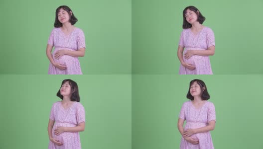 摄影棚拍摄美丽的亚洲怀孕妇女闭上眼睛白日做梦在绿色的摄影棚背景高清在线视频素材下载