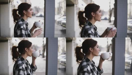 漂亮的女孩站在窗边看风景。用白色的杯子喝茶。侧视图。模糊的背景高清在线视频素材下载