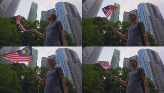 一名年轻男子手持智能手机挥舞着以摩天大楼为背景的马来西亚国旗的慢镜头。去马来西亚旅游的概念高清在线视频素材下载