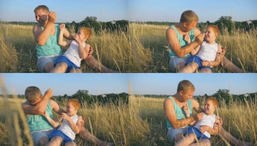 父亲在室外给小儿子挠痒痒。一个快乐的年轻人坐在草地上和他的宝贝儿子玩。快乐的爸爸和他的孩子一起在草地上玩。慢镜头高清在线视频素材下载