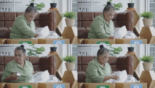 亚洲老年妇女坐在家里分类废物，如玻璃瓶，塑料瓶，金属材料要扔掉。把废物分类再循环再造。保护环境高清在线视频素材下载