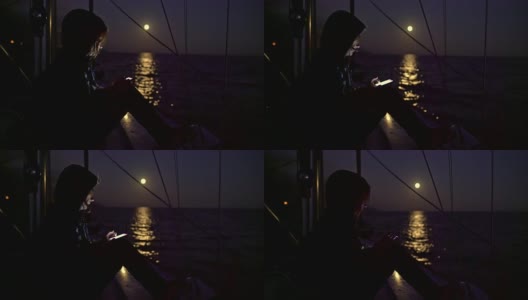 在宁静的满月照耀海洋的夜晚，4K女人在帆船上使用智能手机，实时高清在线视频素材下载