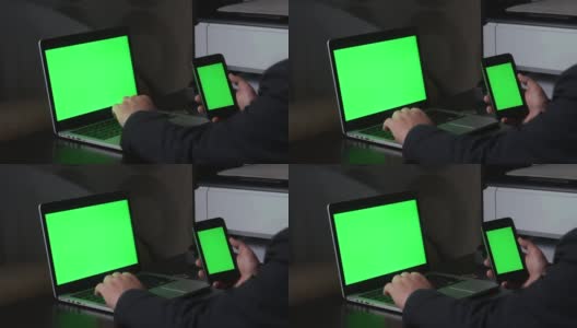 一名男子在一台绿色屏幕的笔记本电脑前工作。他手里拿着一部绿色屏幕的智能手机。附近有一枚加密货币。Chromakey高清在线视频素材下载