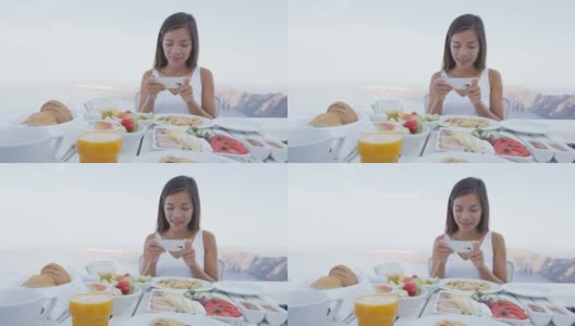 美女用手机app吃早餐。餐桌上有各种各样的健康食品。女游客正在圣托里尼岛的露台度假胜地放松。高清在线视频素材下载