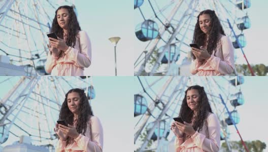 一个长发女孩站在大摩天轮和游乐场附近使用智能手机。慢动作高清在线视频素材下载