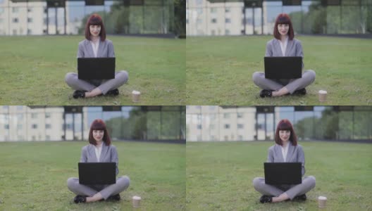 穿着正装的年轻女子，盘腿坐在绿草地上，在办公室外使用笔记本电脑高清在线视频素材下载