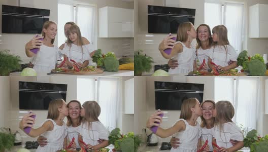 两个可爱的姐妹在厨房做饭的时候亲吻她们心爱的迷人的母亲。女孩们穿着白色衣服亲吻妈妈，在家里厨房用智能手机自拍高清在线视频素材下载