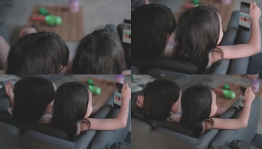 两个亚裔(泰国)黑头发的女孩姐妹坐在房子客厅的灰色沙发上。ฺ两人都在看手机里的视频剪辑。高清在线视频素材下载