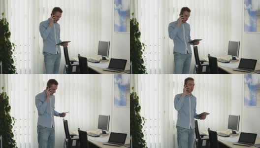 年轻英俊的男子用智能手机打电话，在办公室用平板电脑工作，看着窗外高清在线视频素材下载