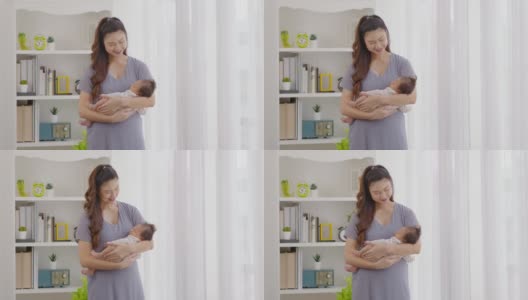 美丽的亚洲妇女抱着新生的婴儿在她的手臂站在舒适的家窗前。快乐的婴儿睡在母亲的怀里，安全舒适。母婴概念高清在线视频素材下载