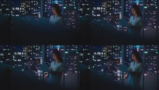 金融分析师使用笔记本电脑，站在窗户旁边与美丽的夜晚城市天际线与摩天大楼。自信的亚洲女商人在现代公司办公室工作。高清在线视频素材下载